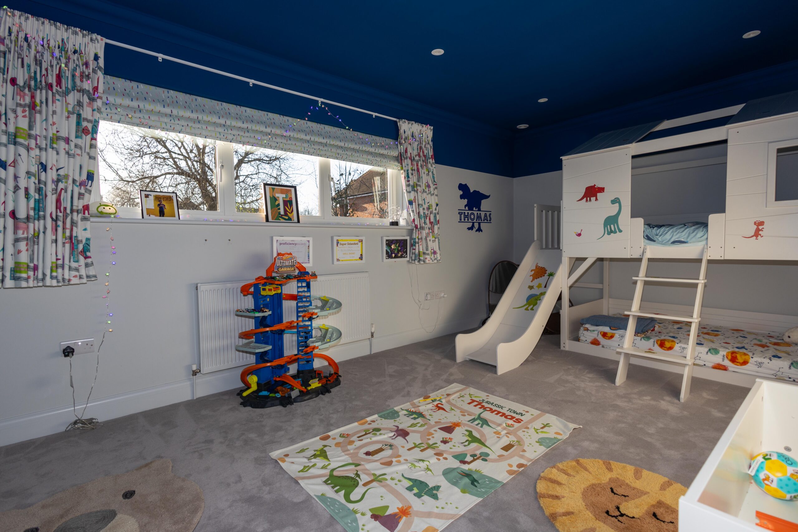 Children's bedroom featuring white bunk beds, a slide, colourful dinosaur decorations and a large window at Haven Lodge, St Albans.